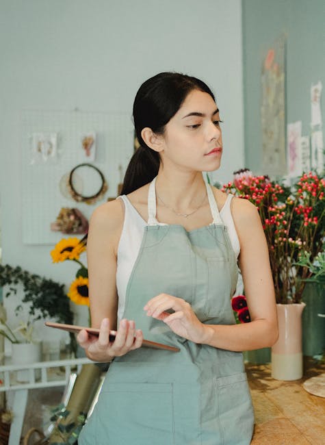 A young woman with dark hair pulled back, wearing a light grey apron over a white sleeveless top, stands in a bright flower shop holding a smartphone in her right hand. She is surrounded by vibrant fl