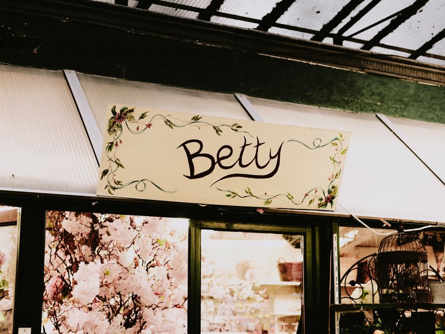 Close-up of a floral shop sign reading 'Betty' with decorative green vines and small pink flowers painted around the border, positioned above a display window filled with pink cherry blossom-themed de