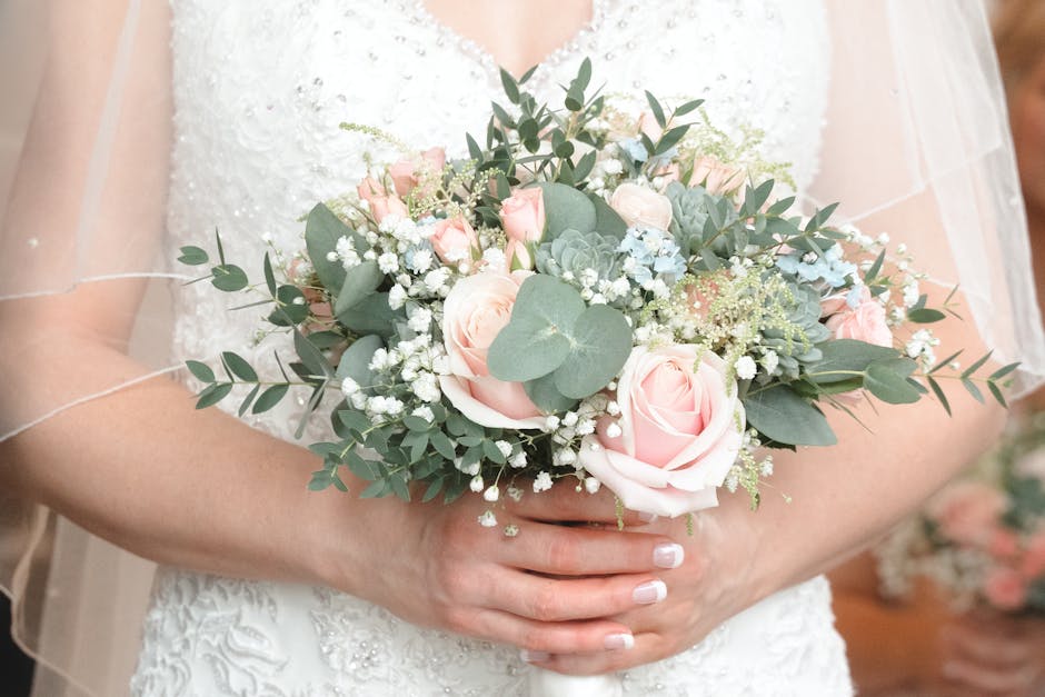 A bride dressed in a white lace wedding gown holds a floral bouquet featuring soft pink roses, white gypsophila, and pale blue hydrangeas, accented with eucalyptus leaves and silver dollar eucalyptus,