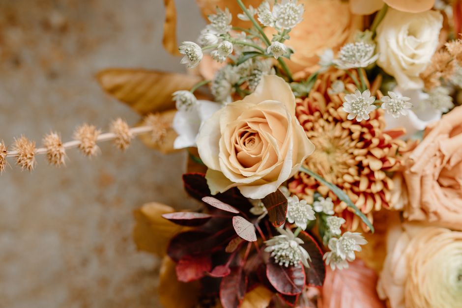 A close-up of a floral arrangement featuring a cream-colored rose with delicate, softly curled petals, surrounded by small white filler flowers and peach-hued carnations. The bouquet includes dark red
