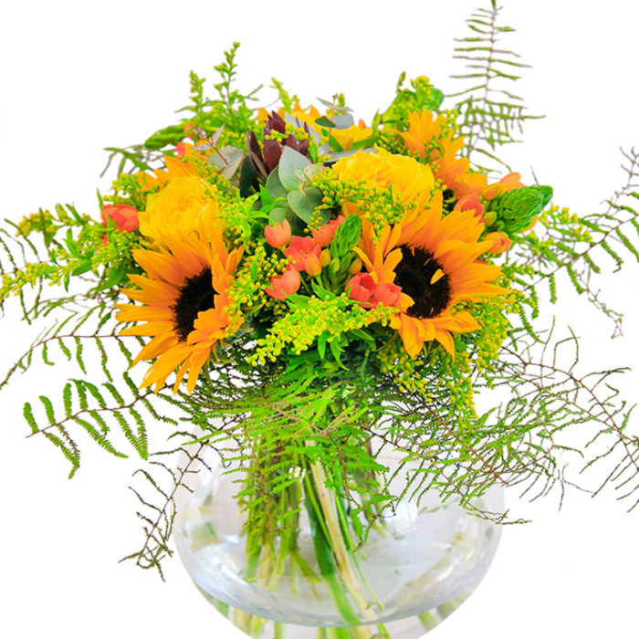 The first impression of this Colney Hatch bouquet is sunshine distilled-a lively tableau where radiant sunflowers and golden yellow roses beam together, their petals unfurling in generous layers that almost seem to invite the warmth of the season indoors. Each sunflower stands tall, its center a dark velvet ringed by a halo of vivid petals, while the roses display spirals of lemony softness, their scent echoing faintly of fresh mornings in Alexandra Park. Between these main blooms, clusters of glossy scarlet Hypericum berries catch the eye, their bright red hue sparkling against a backdrop of lush green foliage. Airy fronds of fern create a sense of lightness, gently cascading outward like the verdant canopies seen in local gardens, and slender spikes of golden filler provide subtle transitions between hues. Silver-tinged eucalyptus leaves peek from the depths of the arrangement, rounding out the natural woodland feel. Cradled in a clear, spherical vase reminiscent of a bubbling spring in Muswell Hill, each stem is visible-every twist and angle telling a story of balance and care. Illuminated by soft daylight, the bouquet radiates happiness and hope, making it perfect for celebrating anniversaries, brightening birthdays, or simply lifting spirits on a grey North London afternoon. Created with pride by Colney Hatch Flowers, this arrangement is more than a display-it's an invitation to pause and find joy in the everyday beauty of our local landscape.