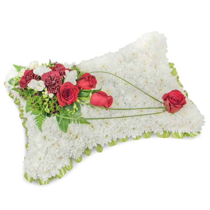 An exquisitely made rectangular memorial cushion, its surface a sea of pristine white chrysanthemums packed so tightly they form a soft, tactile blanket that suggests rest and tenderness. The pillow's edges are finished with a whisper of light green ribbon or fine foliage, just visible beneath the crown of flowers to lend a subtle outline and a hand-finished touch. Arranged on the upper left corner is a vibrant cluster of deep red roses and dark burgundy carnations; the roses range from tight buds to fully opened blooms, their velvety petals providing a rich counterpoint to the pale chrysanthemum field. Interspersed among them are small white accent blooms-likely freesias or spray chrysanthemums-that lift the cluster with delicate highlights. Behind the blooms, lush greenery including fine fern fronds and broader leaves gives structure and an organic backdrop, while long, graceful strands of bright green bear-grass or similar foliage arc across the pillow toward the lower right. These sweeping lines create a sense of movement and connection, culminating in a solitary red rose that rests gently on the lower right area, held visually by the curving foliage and symbolising devotion and remembrance. The overall impression is one of respectful, understated beauty: a funeral pillow crafted for quiet reflection and comfort, suitable for services in and around Colney Hatch, whether laid at a chapel near Coppetts Wood or sent to a family home in New Southgate. The arrangement's textures, colours and scent convey a heartfelt tribute.
