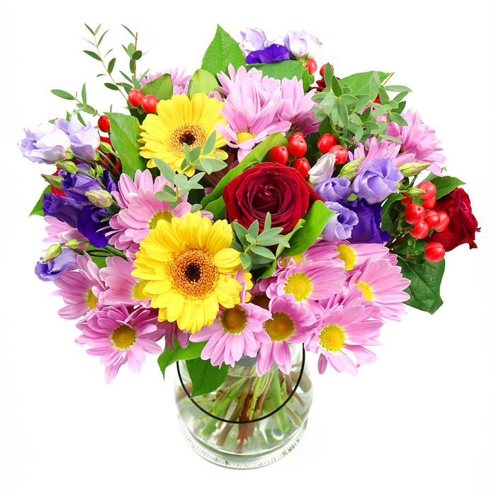A striking hand-crafted bouquet overflows from a cylindrical clear glass vase, positioned against a bright white background that highlights every detail. At its heart, richly coloured red roses unfurl in layered spirals, the velvety petals suggesting timeless affection and sophistication. Nestled close by, two yellow gerbera daisies beam their cheerful faces, dark brown-orange centres ringed with vibrant golden petals that add a burst of sunshine energy. Encircling these central blooms, a generous collar of pink daisies or chrysanthemums spreads outward, each little flower marked by feathery petals and a soft yellow core, creating a cushion of gentle pastel colour. Slender stems of purple and lavender lisianthus rise among the daisies, their bell-shaped flowers and softly curling edges introducing cooler, more serene tones that balance the warmth of the roses and gerberas. Between petals and leaves, clusters of smooth red hypericum berries appear as tiny round accents, lending the design playful texture and a contemporary feel. Layers of foliage in varied greens-some leaves broad and glossy, others thin and airy-frame the flowers and give the arrangement a lush, natural silhouette, reminiscent of a walk through a leafy corner of Colney Hatch Park. Through the transparent vase, you can see the carefully trimmed stems arranged in clear water, a quiet sign of professional attention and freshness. The bouquet feels joyful yet composed, perfect for a birthday, a heartfelt thank-you, or simply to bring colour and comfort into a home near Colney Hatch, Bounds Green, or the nearby residential streets on a calm afternoon.
